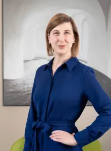 Das Portraitfoto von Henrike Koch. Frau Koch hat glatte, mittellange dunkelblonde Haare, trägt ein blaues Kleid und lächelt den Betrachter mit gespitzem Mund humorvoll an. Im Hintergrund hängt das großformatige Foto eines lichtdurchfluteten Kirchenraums.